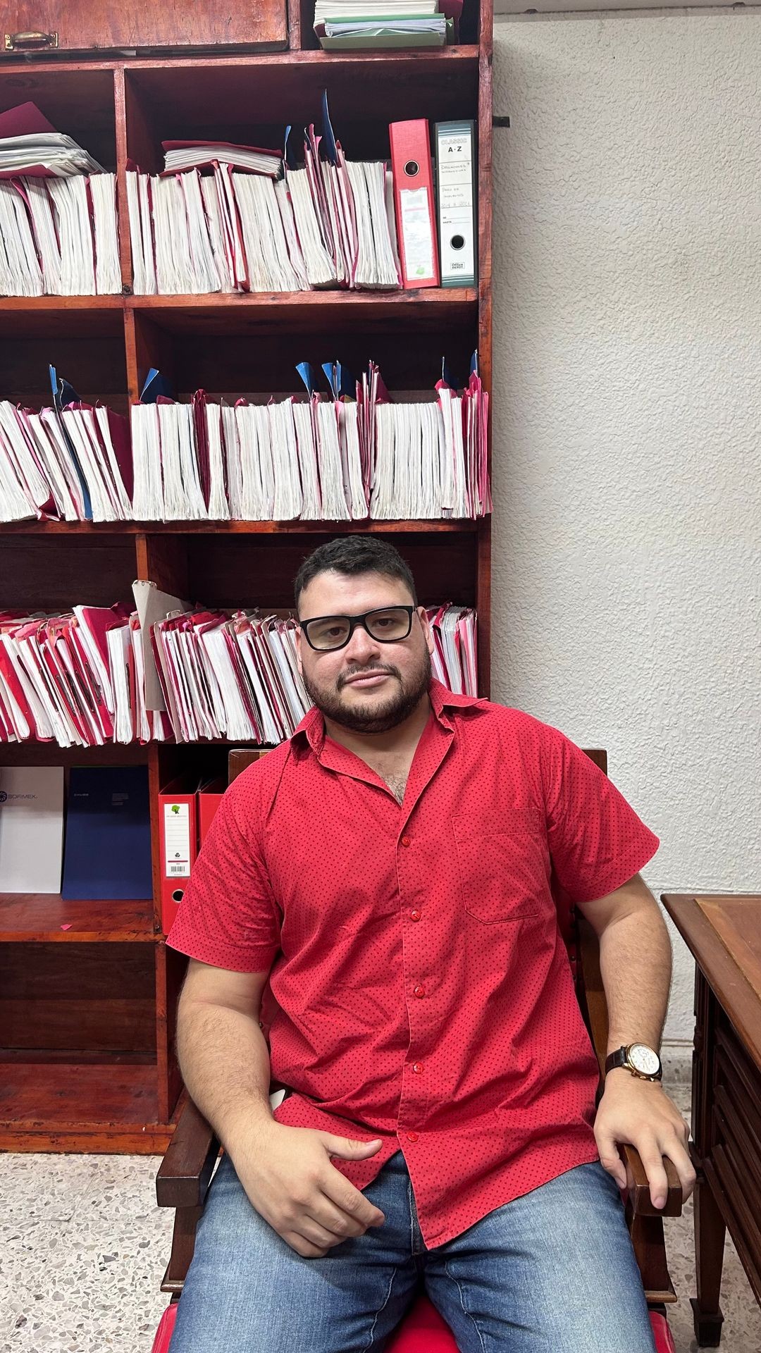 Man in a red shirt sitting in front of a bookshelf filled with organized files and folders.